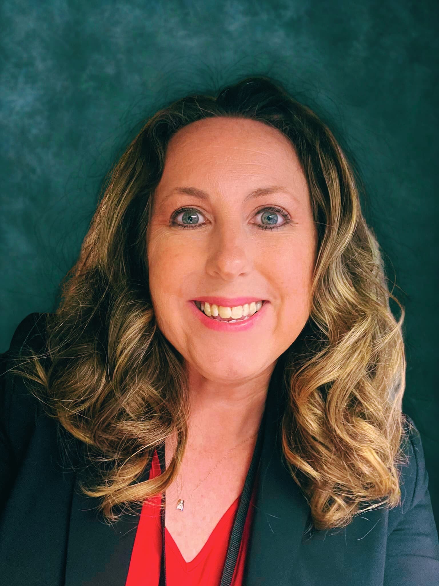 Smiling woman with wavy hair wearing a black blazer and red top, green background.